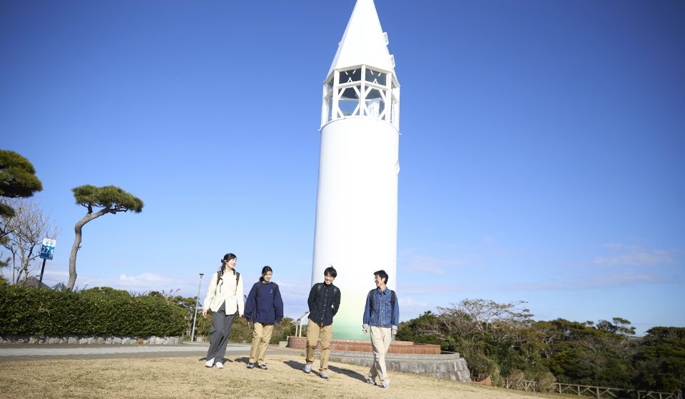 [写真] 青空の下、白い灯台の前を歩くグループの様子
