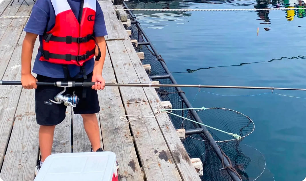 [写真] ライフジャケットを着用し、桟橋の上で真剣に釣り糸を垂らす子供の様子