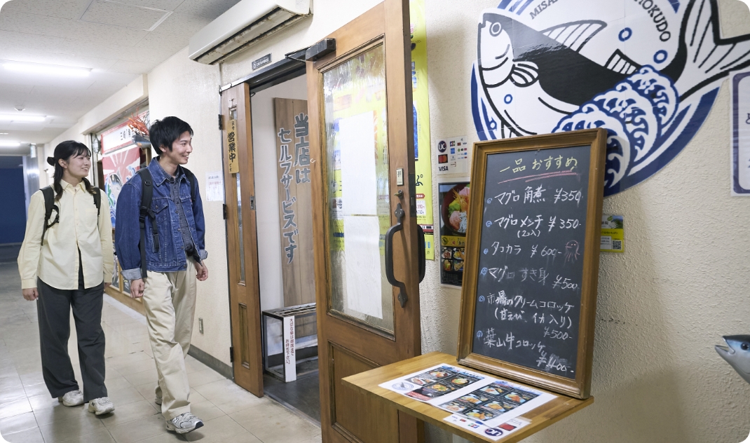 [写真] 「三崎食堂」の入り口で、おすすめメニューが書かれた黒板を眺めながら楽しそうに歩く二人