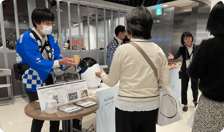 [写真] 法被姿のスタッフが来場者に試供品を渡している様