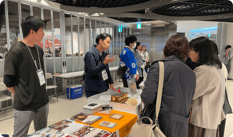 [写真] 特産品の販売ブースで来場者に説明をするスタッフ