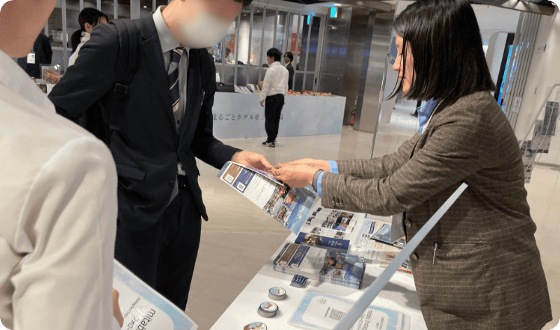 [写真] パンフレットを渡しながら来場者に対応するブースのスタッフ