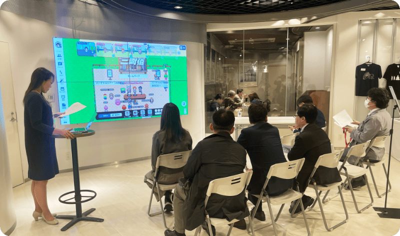 [写真] 大画面モニターの前でプレゼンテーションを行うスタッフと聴講者たち